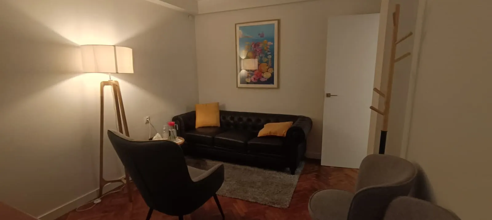 La misma consulta vista desde la entrada: sofá de piel negro con cojines amarillos, butacas, lámpara de pie encendida y un cuadro de colores en la pared.