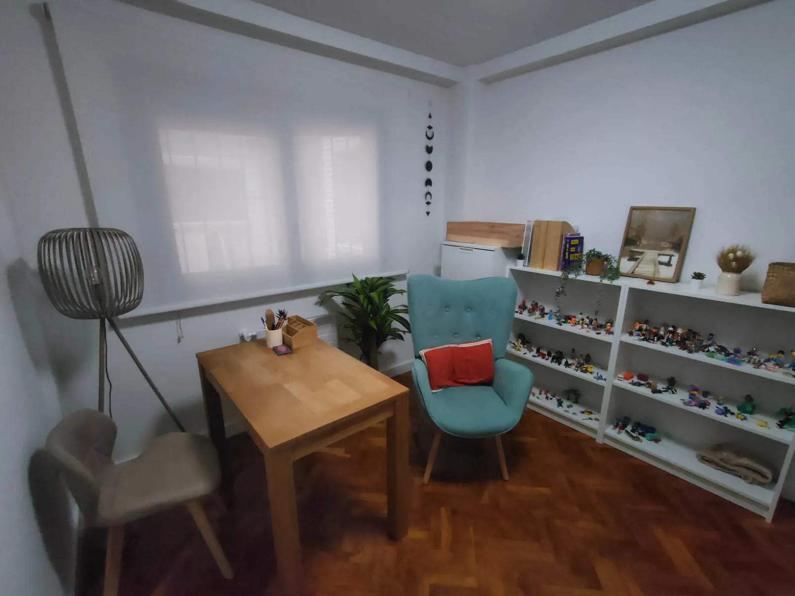 Despacho 1 con mesa de madera y lápices, butaca turquesa, lámpara de pie y estanterías con figuras de juego.